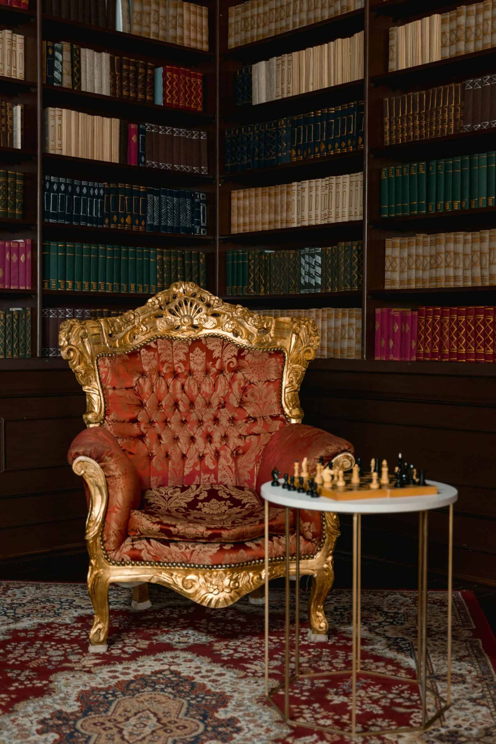 Elegante silla de estilo vintage con tapizado en tonos rojos y dorados, en una biblioteca con estanterías llenas de libros antiguos y una alfombra tradicional, creando un ambiente sofisticado y acogedor.